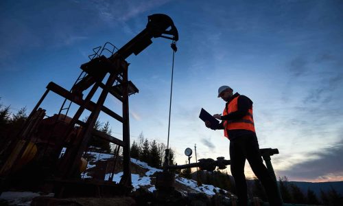 Low angle of petroleum engineer in work vest looking at oil pump rocker-machine and writing on clipboard. Oil worker checking work of balanced beam petroleum pump jack under beautiful evening sky.