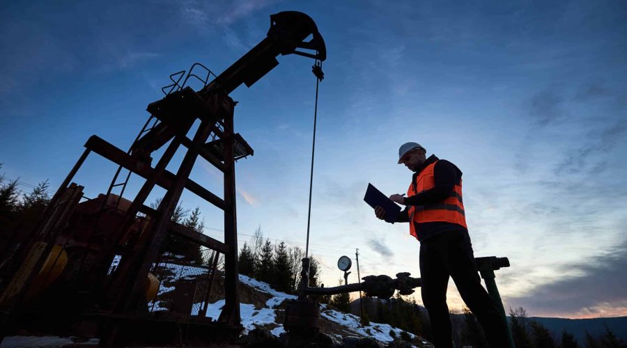 Low angle of petroleum engineer in work vest looking at oil pump rocker-machine and writing on clipboard. Oil worker checking work of balanced beam petroleum pump jack under beautiful evening sky.